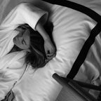 Person lying on a bed with a blanket and pillow, wearing a coat.