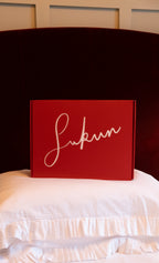 Red box with 'sukun' written on it in white cursive text, placed on a white surface with a dark headboard in the background.