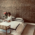 Woman sitting on a bed with white bedding against a brick wall